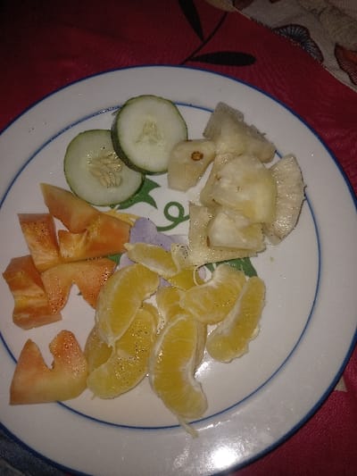 A white ceramic plate, edged with a thin blue line and featuring a faint green and purple floral pattern in its center, holds an assortment of prepared fruits and vegetables. On the upper left, two circular slices of green cucumber are visible, one displaying its inner seeds. To the upper right, multiple irregular, light yellow-white chunks, consistent with peeled pineapple or yam, are arranged. The lower left quadrant contains several wedge-shaped pieces of a reddish-orange fruit, possibly tomato or papaya, some exhibiting lighter streaks. In the lower-center to lower-right, numerous peeled, individual segments of an orange or tangerine are present. Fine, dark specks are scattered across the surfaces of the orange segments and other items. The plate rests on a red fabric surface, patterned with dark, indistinct floral or leaf motifs. Part of a lighter, patterned fabric is partially visible to the right. The scene is illuminated by ambient, possibly artificial, light, creating some glare on the plate's surface. This scene is located in Igbede, Nigeria. No individuals are depicted, and no active interactions are occurring.