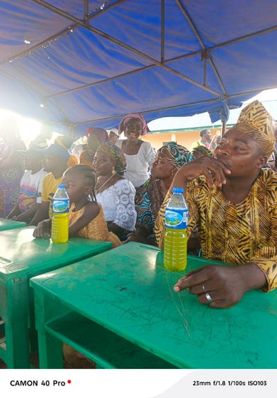 The image depicts a group of people seated at green tables under a blue canopy, likely at an event in Gidan Kogi, Nigeria. A man in a yellow patterned robe and matching cap is in the foreground, his hand near his mouth with a silver ring on his finger. A bottle of "Swan" water sits on the table in front of him. To his left, a woman wears a colorful head wrap, and a younger girl with braided hair sits at the table. Behind them, other people are visible, including a woman in a white lace blouse and purple head wrap, who is smiling. The canopy above is supported by metal beams. Sunlight filters through the left side of the frame.