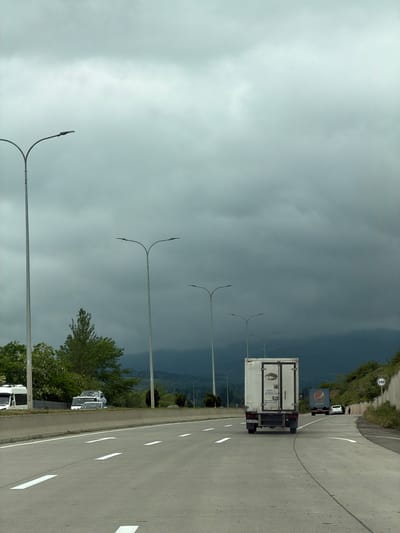The image depicts a highway in Tserovani, Georgia under a stormy sky. A white delivery truck with visible license plate "AA-748-GI" is traveling on the right lane. Further ahead, a Pepsi truck and another white vehicle are also on the road. The highway has white lane markings. Street lights line the side of the road. Trees and vegetation are visible along the roadside. A speed limit sign (110) is present on the upper right side of the image.