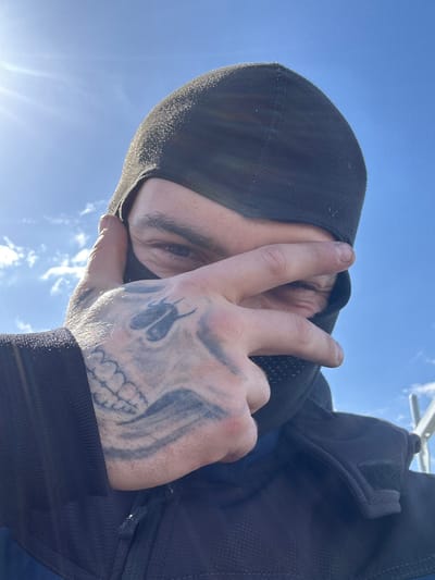 A man is pictured from a low angle, likely a selfie, against a bright sky with wispy clouds. He is wearing a dark hooded garment, possibly a balaclava or ski mask, that covers most of his head. Only a portion of his face, including an eye and part of his brow, is visible. His hand is raised, forming a "peace" or "victory" sign with his index and middle fingers. The hand has a prominent tattoo depicting a skull and other shapes. He is also wearing a dark-colored jacket. The location is not specified and Roßdorf, Germany, is not mentioned in the image or its description.