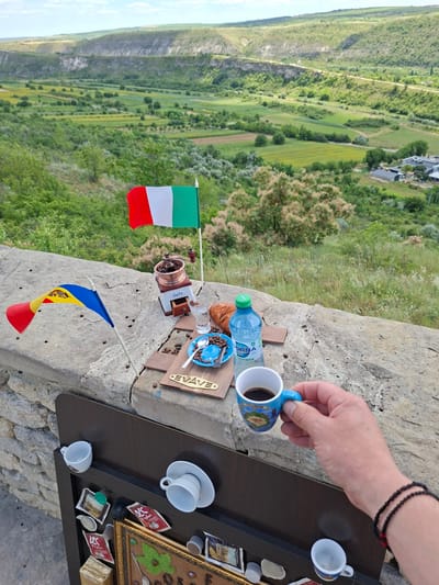 In Trebujeni, Moldova, a scenic vista unfolds with rolling green hills and a valley. Perched on a stone wall, a small display is set up with two miniature flags, one Italian and one Moldovan. A vintage coffee grinder, a glass of water, a croissant, and a blue plate with coffee beans and a spoon are arranged on a small wooden platform. A bottle of water and a miniature cup of coffee are also present. Below the wall, a dark panel displays several coffee-related items including small cups, sachets, and capsules. A hand holding a cup of coffee is in the foreground, obscuring some of the view.