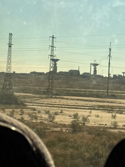 The image depicts a wide, arid landscape under a pale blue sky, characteristic of Toretam, Kazakhstan. In the mid-ground, several large, lattice-style electricity pylons stand tall, supporting power lines that stretch across the horizon. Behind the pylons, a collection of buildings and structures are visible on a raised earthen berm, suggesting an industrial or research facility. Prominently featured among these structures are several large, dish-like antennas, indicative of a telecommunications or space tracking station. The foreground is dominated by a section of what appears to be a vehicle's interior, possibly a car or bus, with a dark, curved frame partially obscuring the view. The landscape itself is sparsely vegetated with dry grasses and low bushes, interspersed with barren, sandy patches. The overall impression is one of a functional, possibly remote, technological site situated within a vast, dry expanse.