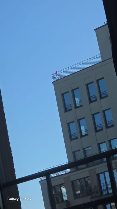 A photograph taken outdoors shows a portion of a light-colored, multi-story building against a clear blue sky. The building has a modern design with numerous rectangular windows, many of which appear to reflect the sky. On the roof of the building, two small, red lights are visible. In the foreground, a dark metal railing runs horizontally across the frame, partially obscuring the view of another building. The bottom left corner of the image displays the text "Galaxy Z Fold7".