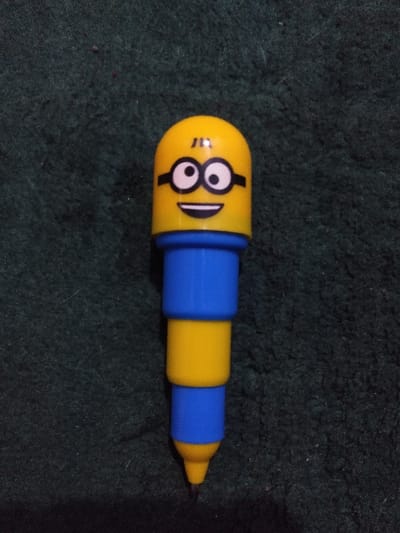 A single Minion-themed writing instrument, primarily yellow and blue, is positioned centrally against a dark green textured background. The top segment is a yellow, rounded cap depicting a Minion's face, characterized by black-rimmed goggles with white eyes and black pupils, and a black smiling mouth. The body of the instrument consists of alternating blue and yellow cylindrical segments. It terminates in a yellow conical tip from which a dark, fine writing point extends. The background is a uniform dark green, appearing to be a coarse textile surface such as carpet or shag fabric, exhibiting slight variations in texture and minor surface debris. No individuals are present, and no actions or interactions are depicted. The specific location within Ciudad de San José, Costa Rica, is not discernible from the visual information provided.