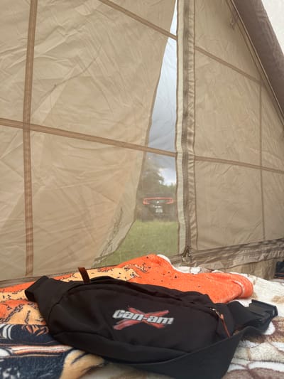 The image shows the interior of a tent with clear plastic windows. Through the windows, an orange object, possibly a vehicle, is visible in an outdoor setting with green grass and trees. On the right side of the frame, resting on a patterned blanket, is a black fanny pack with a "Can-Am" logo. The tent fabric is a light tan color and appears slightly wrinkled. Zippers and seams are visible on the tent material. The lighting suggests it is daytime.
