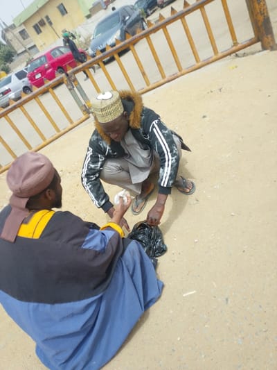 An outdoor scene in Daurawa, Nigeria, depicts two adult males on an unpaved, light brown ground surface. In the foreground, one male is seated, viewed from behind and slightly to his left. He wears a dark blue, black, and yellow long garment and a brown head covering. Facing him, another male crouches, wearing a black jacket with white stripes on the sleeves, a light grey tunic, tan trousers, and a patterned cap. The crouching male extends a small, wrapped white object in his right hand towards the seated individual, while his left hand rests on a black plastic bag on the ground between them. A horizontal orange metal barrier runs across the mid-ground, separating the immediate interaction from a paved road. Beyond the barrier, multiple vehicles are parked, including a red hatchback, a grey sedan, and several dark-colored sedans. A third individual in dark clothing is visible walking in the background. Buildings are partially visible in the far background.