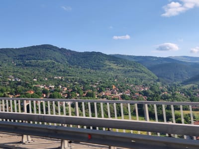This is a landscape photograph taken from a bridge, looking out over the town of Eleshnitsa, Bulgaria. The foreground is dominated by the bridge railing, a grey metal structure with vertical bars. Beyond the bridge, the scene opens up to a vista of rolling, tree-covered hills. Nestled within the hills is the town of Eleshnitsa, characterized by numerous houses with red-tiled roofs. The houses are scattered across the slopes, interspersed with abundant green trees. The sky above is a clear, bright blue with a few wispy white clouds. The overall impression is one of a rural, scenic setting.
