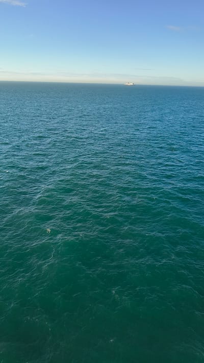 The image shows a wide expanse of the ocean under a clear blue sky. A ferry is visible in the distance, traversing the horizon. The water is a deep teal color, with gentle waves rippling across its surface. A faint line of clouds hugs the horizon. The scene appears to be a calm day at sea, with no discernible urban landscape or human activity other than the ferry. The location is unknown, but it could be a coastal area in the United Kingdom, given the presence of a ferry.