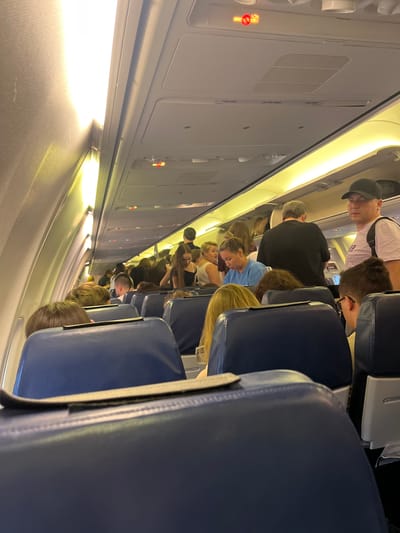 The image depicts the interior of an airplane cabin, viewed from a low angle looking down the aisle. Several passengers are visible, seated and in motion within the narrow confines of the aircraft. The seats appear to be upholstered in dark blue fabric. The overhead compartments and interior wall panels are a neutral gray or white. The lighting is a warm yellow, suggesting either the cabin lights are on or it is not daytime, or perhaps it is a time of day when artificial lighting is favored.

In the foreground, the heads and shoulders of two passengers are visible, their hair color ranging from blonde to dark brown. Further down the aisle, a line of passengers are seen standing and moving, indicating boarding or disembarking. A man wearing a black baseball cap and a white t-shirt is prominent in the upper portion of the image. Another man in a black t-shirt is visible behind him. A woman with blonde hair in a light blue shirt is also discernible among the passengers.

Notable details include a red illuminated sign with a symbol of a cigarette crossed out, indicating a "no smoking" rule. There is also a vent or speaker grille on the wall panel. The overall atmosphere suggests a typical flight experience, with people navigating the aisle and settling into their seats. There are no direct cues for time of day, weather, or specific emotions. The location context of Stolbishche, Russia, cannot be confirmed from the visual information within the image.
