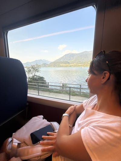 A woman with dark hair, wearing a white t-shirt and a watch, looks out the window of a train at a lake and mountains. Her arms are crossed, and a pink bag with a smartphone is on her lap. The scene is set in Calceranica al Lago, Italy, with a clear sky and a calm lake visible.