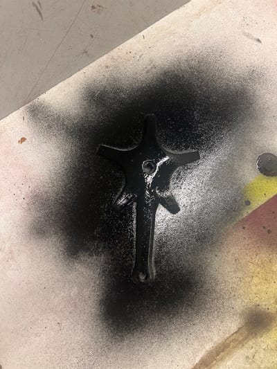 A black, star-shaped metal object with one longer arm rests on a concrete surface. It appears freshly spray-painted, with overspray creating a diffused, dark aura around the object. A small circular hole is visible in the center of the star, and another hole is present at the end of the longer arm. Discoloration and other materials, like paint, are noticeable in the background.