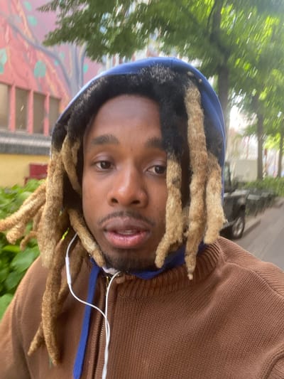A selfie shows a Black man with tan dreadlocks, wearing a brown zip-up hoodie with a blue hood. He has a small mustache and beard. White earphones hang from under his hood. The background appears to be an outdoor urban environment, possibly in Le Pré-Saint-Gervais, France, with blurred buildings and greenery visible. A colorful mural is partially seen in the background.