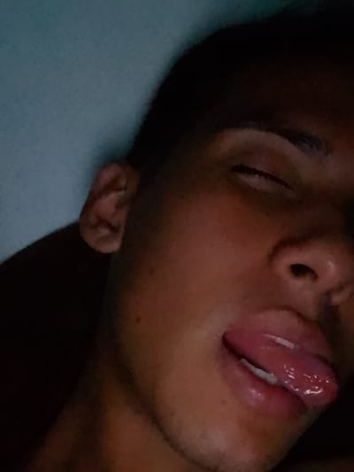 The image is a close-up portrait of a person, likely a young adult male, captured from a slightly upward angle. The individual appears to be lying down, possibly in a bed, with their head resting against a soft, light blue background that resembles a pillow or bedsheet.
The lighting is dim, suggesting an indoor setting during nighttime or in a dark room. The subject has dark skin, dark hair visible at the top of the frame and around the ear, and a visible eyebrow. Their eyes are mostly closed, with the whites showing in the upper portion, which could indicate a state of sleep, deep relaxation, or a playful expression. Faint stubble is visible around the chin and jawline.
The most prominent feature is the person's mouth, which is slightly open with their tongue extended outwards. The lips are full and appear glossy, and the tongue is visibly wet and shiny. This expression could be interpreted as playful, relaxed, or a candid moment. The overall mood of the image is intimate and casual. No text is visible in the image.