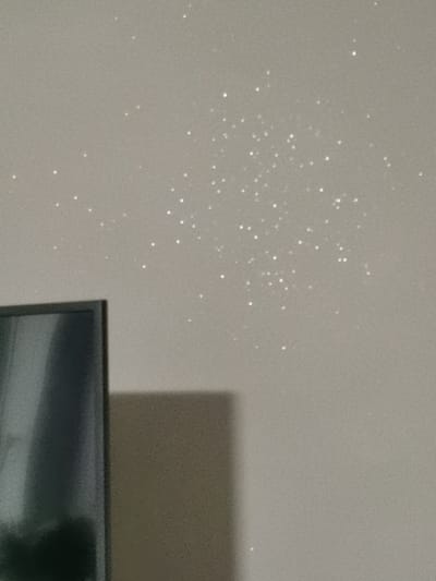 The image depicts a close-up, eye-level, medium shot of a wall with small, shimmering particles scattered across its surface. The particles appear to be reflective and are distributed in a somewhat clustered pattern. In the lower left portion of the image, a dark, rectangular object, likely a television screen, is partially visible. The wall itself is a light, neutral color, possibly beige or gray, with a slightly textured finish. The lighting suggests an indoor environment, with a soft, diffused light source casting a subtle shadow from the television. No people or specific actions are depicted. The location of Bourg-lès-Valence, France, is not identifiable from the visual information present in this image.