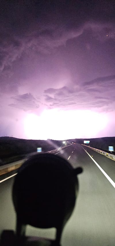 The image depicts a highway at night with a purple sky illuminated by a distant lightning strike. In the foreground, a dark, out-of-focus object, possibly the interior of a vehicle, obscures the lower portion of the view. The highway itself is well-lit, with white lines marking the lanes and reflectors lining the sides. On the left side of the highway, a dark, undulating landscape stretches into the distance, dotted with what appear to be wind turbines on a hill. On the right side of the highway, similar landscape features are visible, along with roadside lighting. The overall scene suggests a stormy night on a rural highway in the vicinity of Malpartida de Plasencia, Spain.