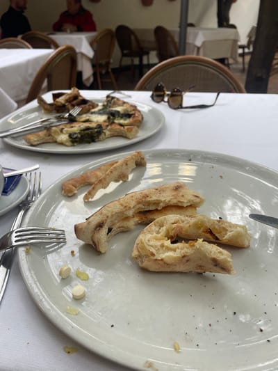 The image depicts an outdoor dining scene at a restaurant in Sorrento, Italy. In the immediate foreground, a light-colored circular plate rests on a white tablecloth, containing several pieces of partially consumed, light-brown bread or pizza crust, and two small, white, oval-shaped pills. Scattered food debris, including small yellow fragments and dark specks, are visible on the plate. To the left of this plate, a silver four-tine fork with residue is present, along with the edge of a blue and white patterned cup or saucer. A silver knife handle is partially visible on the right edge of the plate.
In the midground, a second, oval-shaped, light-colored plate is on a white tablecloth. This plate holds remnants of a pizza-like dish, featuring melted white cheese and dark green leafy vegetables, likely spinach, on a light brown crust. A silver fork and knife are positioned on this plate. Behind this plate, a pair of brown tortoise-shell patterned sunglasses with dark lenses rests on the white tablecloth. A wooden chair back with light upholstery is partially visible behind the sunglasses.
The background shows additional tables covered with white tablecloths and accompanying chairs, indicating a bustling restaurant environment. Several blurred human figures are discernible: one in a dark top on the far left, another in a red jacket seated at a table in the center-left, and a standing figure partially visible on the far right. Architectural details include a light-colored wall with terracotta pots mounted on it and a dark vertical pole or tree trunk on the far right. The scene suggests a meal that has been partially or mostly consumed.