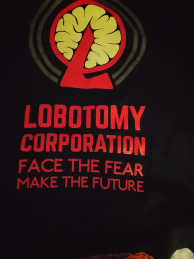 The image shows a black t-shirt with red lettering and a graphic design. The graphic at the top of the shirt depicts a stylized brain with concentric circles behind it, and a large red "L" shape superimposed over the brain. Below the graphic, in red capital letters, are the words:
LOBOTOMY
CORPORATION
FACE THE FEAR
MAKE THE FUTURE
The image was taken indoors and does not provide any information about the location in the city of Zhlobin, Belarus.
