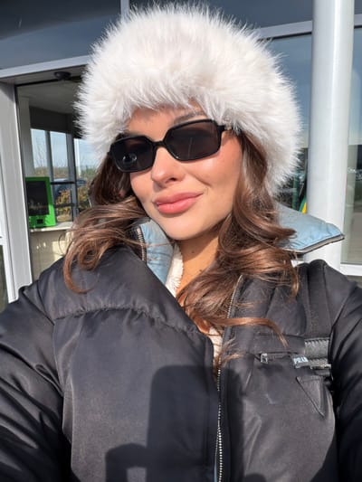 A female individual is depicted in a close-up, likely selfie-style, outdoor photograph. She is wearing a prominent white, fluffy faux fur hat that covers most of her head, dark rectangular sunglasses, and a black puffer jacket with a visible silver zipper. Beneath the puffer jacket, a light blue collar and a cream-colored, textured lining are discernible. A black strap from a bag or backpack is visible over her right shoulder, with a black triangular patch on the jacket bearing the "PRADA" logo. The individual has medium-brown, wavy hair and a subtle, closed-lip smile. Strong sunlight illuminates the scene, casting shadows. In the background, visible through large glass windows, are architectural elements characteristic of a modern facility, including white support columns and a bright green electronic kiosk, possibly a parking payment machine. The probable location, given the city of Colombier-Saugnieu, France, and the visual cues, is Lyon-Saint Exupéry Airport.
