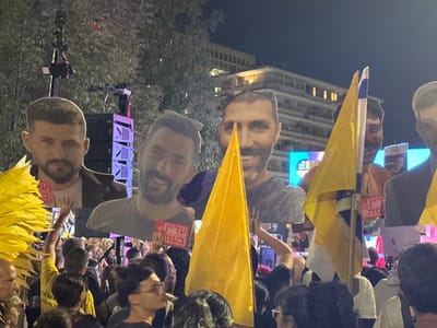 A nighttime outdoor gathering is depicted in Paris Square (Kikar Paris) in West Jerusalem, Israel. A dense crowd of people occupies the foreground and midground, facing forward. Multiple large cardboard cutouts of men's faces are held aloft above the crowd. On the far left, a cutout shows a man with a dark beard, short dark hair, and a serious expression, wearing a dark jacket. A red sign held below this cutout displays partially visible Hebrew text, including "חיים בעזה" (Chaim in Gaza), identifying the individual as a hostage. To his right, a cutout features a smiling man with a dark beard, short dark hair, wearing a grey t-shirt and sunglasses pushed onto his head. Below this, a pink sign in Hebrew reads "עת עדין בנצור" (Adin is still in captivity), indicating another hostage. Further to the right, another smiling man's cutout is partially obscured by a bright yellow flag, a symbol of the "Bring Them Home Now" campaign. Numerous bright yellow flags are visible throughout the crowd, some on poles. An Israeli national flag (blue Magen David on white) is partially visible. In the background, tall speaker stacks and camera equipment are mounted on scaffolding, suggesting a stage or public address system. A large digital screen in the distant background displays illuminated blue and white graphics. Multi-story buildings with illuminated windows are visible behind sparse tree foliage, indicating an urban setting. People in the crowd are primarily seen from the back or side, standing and holding objects. The scene is illuminated by artificial lights emanating from the stage, screen, and surrounding buildings.
