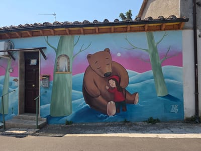This is a photograph of a mural painted on the side of a building. The mural depicts a large brown bear hugging a young child wearing a red hat and jacket. The background of the mural is a landscape with stylized trees and rolling blue hills against a pink and blue sky, suggesting a sunset or sunrise. A small white moon is visible in the sky.
On the left side of the mural, next to the entrance of a building, there is a small niche with a statue of the Virgin Mary. The building's entrance is a dark brown door with a doorknob, accessed by a short set of concrete steps with green railings. Above the door, an air conditioning unit is mounted. There are also mailboxes and a sign on the wall.
To the right of the mural, a white downspout runs vertically along the building's exterior. A small electrical panel with a drawing of an open book and faces is visible on the blue painted wall at the base of the mural.
The scene appears peaceful and comforting, with the bear and child embracing. The overall mood is serene. The image is taken outdoors on a clear, sunny day. The building's tiled roof and the style of the architecture, along with the "Viterbo, Italy" context, suggest this is a residential or small business building in a European setting. The mural is vibrant and colorful, indicating it is a focal point of the building's facade. The visible text includes a sign on the door that says "Vende" (Italian for "For Sale") and "A.C.A.S." on the electrical panel. There is also illegible text on the downspout.