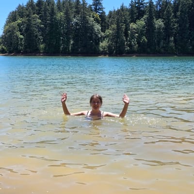 In Cherry Grove, United States, a young woman is swimming in a lake. She is partially submerged in the water, with her upper body visible. Her arms are raised in the air, and she is smiling at the camera. The water is clear and calm, with sunlight reflecting off its surface. In the background, a dense forest lines the edge of the lake. The sky above is a clear blue.