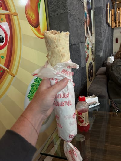 A person's arm and hand are holding a tightly wrapped shawarma in front of a brightly colored mural and a stone wall. The shawarma is wrapped in white paper with red text and a green graphic of lettuce. The mural features a stylized depiction of food items. Behind the shawarma, a dark gray stone wall is visible, adorned with posters. On a glass table in the foreground, a bottle of tomato juice, a small white container with utensils, and sunglasses are placed. The background shows a seating area with plush dark gray couches. This scene appears to be in a casual dining establishment in Tsovagyugh, Armenia.