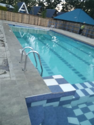 A long, rectangular swimming pool with clear, turquoise water stretches toward the background. White lane markers run along the pool's floor. The pool's edge features gray tiles and a checkerboard pattern of blue and white tiles leads into the water. A metal ladder provides access to the pool. A bamboo fence and several blue-roofed structures are visible in the background under a muted sky.