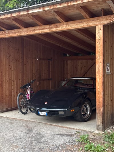 The image depicts a black car parked inside a wooden carport. The car appears to be a vintage model, with its interior visible and a license plate that reads "ZA647VL". To the left of the car, a bicycle with a pink water bottle and a pink bag attached to the frame is parked. The carport is constructed from wooden beams and planks, with a slanted roof. The scene is outdoors, as suggested by the presence of greenery and gravel outside the carport. The lighting suggests it is daytime. A small sign with a red circle and a line through a figure is visible on the wooden ceiling of the carport. The location is identified as La Ila - Stern - La Villa, Italy.