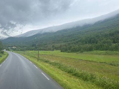 The image shows a road curving to the left, flanked by green grassy fields and a dense forest. In the background, a misty mountain range is visible, with the peaks obscured by clouds. The sky is overcast with a dull gray hue. A white line marking divides the lanes of the asphalt road. There are utility poles along the side of the road. The scene appears to be a rural or semi-rural landscape in Norway.