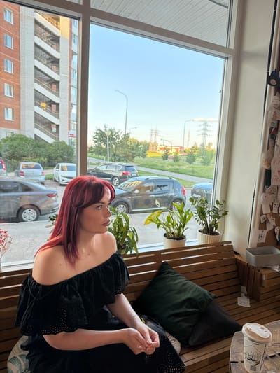 A young woman with bright red hair, wearing an off-the-shoulder black lace top, sits on a wooden bench in a cafe. She is looking out a large window that offers a view of an urban street scene. Outside, cars are parked along the street, and a multi-story brick building with external staircases is visible. The sky is clear and blue, with power lines and trees in the distance. Inside the cafe, several potted plants sit on the windowsill, and a bulletin board covered with papers and drawings is on the right side of the image. A coffee cup is on a table in the foreground. The scene appears to be set in Timofeyevka, Russia.