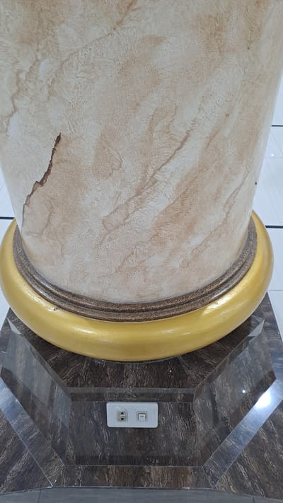The image shows a close-up of a decorative column in what appears to be an indoor setting, possibly a public building or a well-decorated residence. The column is large and cylindrical, with a textured surface that mimics marble or a similar stone. It has a light beige or cream color with swirling patterns of brown and tan. A dark, irregular crack or imperfection is visible on the lower left portion of the column. The column is accented by a thick, golden band that wraps around it, creating a prominent decorative element. This band has a smooth, glossy finish.
Behind the column, the wall features a dark, marble-like pattern with a geometric design. The pattern is made up of angled, beveled surfaces that create a sense of depth and angularity. A white electrical outlet and light switch plate are mounted on this wall, positioned to the right of the column. The outlet has two sockets, and the switch appears to be in the "off" position. The lighting in the scene is bright, suggesting it is daytime or the area is well-lit artificially. There are reflections visible on the patterned wall, indicating a smooth, possibly glossy surface. No people or animals are present in the image, and no specific activity or event is depicted. The overall impression is one of a luxurious or well-appointed interior space with architectural embellishments. The presence of the electrical outlet suggests it is a functional space. There is no visible text in the image. The location context provided is Lhokawe Teungoh, Indonesia, which adds a regional aspect to the description.
