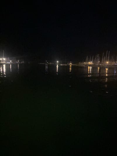 This is a nighttime, eye-level, full shot of a marina in Agde, France. The water in the foreground is dark and reflects the lights of the docked boats. In the background, a dense cluster of sailboats are moored, their masts creating a vertical line against the dark sky. Lights from buildings and street lamps illuminate the marina, casting long reflections on the water. The scene is quiet and still, suggesting a peaceful, serene atmosphere.