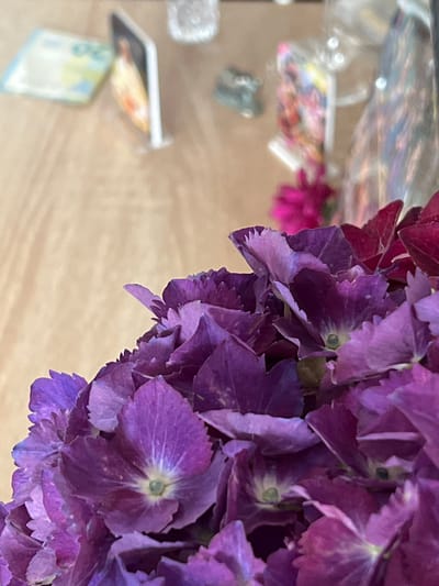 The image shows a close-up of vibrant purple hydrangeas in the foreground, with their delicate petals and intricate centers clearly visible. The background is blurred, but it appears to be a wooden surface with some items placed on it. To the left, a light blue object with some markings, possibly money, is visible. Further back, there are some small, indistinct objects that could be decorative items or photographs. A hint of a bright pink flower can be seen behind the hydrangeas. The overall impression is of a still life arrangement, possibly on a table in a home or shop. The image does not contain any specific elements that would indicate its location in Bunbeg, Ireland.