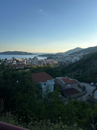 The image shows a panoramic view of the coastal city of Čučuci, Montenegro, at sunset. In the foreground, lush green trees and vegetation frame the scene. Below, two houses with red-tiled roofs are visible, one white and the other a light brown color. A paved road winds past the houses, with two parked cars. The middle ground features a dense cluster of apartment buildings and other structures, some with white facades and others with varying shades of grey and beige. Beyond the city, a body of water, likely the Adriatic Sea, stretches to the horizon, with a distant island in the background. The sky is a clear blue, with a gradient of lighter hues near the horizon, indicating the setting sun. The overall scene depicts a tranquil evening in a coastal town nestled among rolling hills.