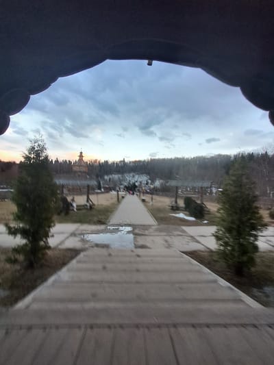 The image shows an exterior view, possibly taken from a slightly elevated position or from within a building, of a scene that includes features consistent with the city of Vzglyadnevo, Russia. The foreground contains a dark, undefined shape on the left, possibly part of clothing. A paved walkway extends into the scene on the right, with puddles suggesting recent rain. Beyond the walkway, there is a grassy area with trees and what appears to be a wooden structure or fence. In the distance, a building with a distinctive tower or spire is visible amidst a line of trees. The sky is cloudy, and the light suggests either dusk or dawn. There are no people visible in the image. The scene's elements include natural elements (trees, sky) and man-made structures, with possible signs of recent precipitation.