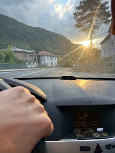 The media file shows a view from inside a car, looking out the windshield. The sun is setting, casting a warm golden light and creating lens flare through the slightly dirty glass. The scene outside depicts a hilly, tree-covered landscape with a few buildings visible on a slope. The perspective is from the driver's seat, with a hand on the steering wheel visible in the foreground. To the right, a car's digital display shows information such as "Olovo" (possibly a location or media source), "Range 462 km", "-Consumption 4.1 Ltr./100 km", and "Inst. consumpt. 0.0 Ltr./100 km". It also displays the time as "0.22:23". Several coins are placed on a textured surface next to the display. The overall impression is of a drive in the countryside during sunset, with a hint of recent rain suggested by the water droplets on the windshield. The presence of the coins and the car's digital display suggests a practical, everyday setting. The low angle of the sun and the warm tones indicate it is late afternoon or early evening.