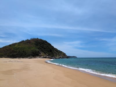 The image depicts a serene beach in Sa Huynh Ward, Vietnam, on a bright, partly cloudy day. The foreground is dominated by a wide expanse of golden sand, gently sloped towards the turquoise ocean. Small waves with white foam are lapping the shore, creating subtle patterns in the wet sand. In the mid-ground, a lush, green, tree-covered hill rises dramatically from the coastline, its slopes dotted with rocky outcrops. The water transitions from a bright turquoise near the shore to a deeper blue towards the horizon. In the far distance, a faint structure, possibly a lighthouse, can be seen on another headland. The sky is a clear blue, streaked with wispy white clouds that add texture to the upper portion of the image. There are no people visible in the scene, contributing to the tranquil and isolated atmosphere of the beach.
