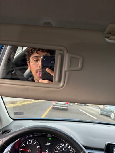 A selfie reflection in a car visor mirror. The man is holding a smartphone, sticking out his tongue. The reflection includes a partial view of the driver's side window, the steering wheel, and the dashboard. Outside the car, a street with other vehicles and lane markings can be seen. The car's speedometer reads 9 km/h, and the odometer is 253 km. The specific location within San Bernardino Chalchihuapan, Mexico is not identifiable from the image.