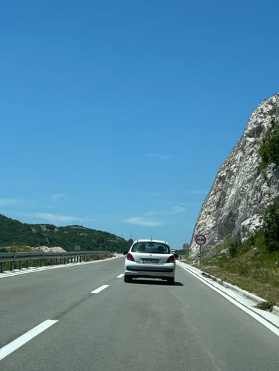 A white hatchback car with license plate "BA BK 765" drives down a paved road on a sunny day. The car is in the center of the frame, traveling away from the viewer. To the right of the road is a steep, rocky cliff face covered in netting. To the left of the road are green, rolling hills. The sky is clear and blue. Two traffic signs are visible on the right side of the road: one indicating a speed limit of 60 km/h, and another prohibiting overtaking. The scene appears to be on a highway outside of a city. The image was taken in or near the city of Obzovica, Montenegro.