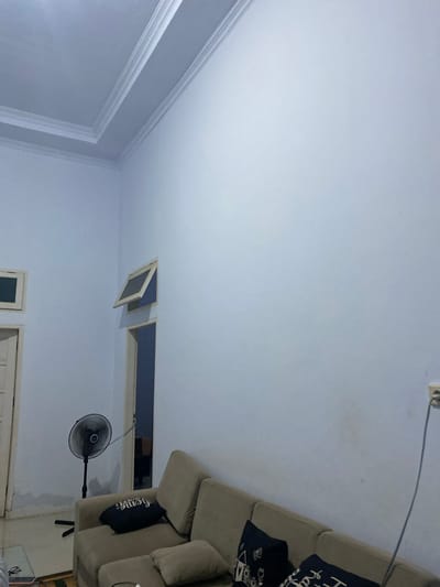 The image depicts an indoor scene, likely a living room, with light blue walls and a white ceiling with decorative molding. On the left side, there is a doorway with a closed door and another small, closed window above it. To the right of the doorway, there is a slightly open ventilation window. In the foreground on the left, a black oscillating fan is standing on the floor. A beige sectional sofa occupies the right side of the image, with several dark-colored throw pillows on it. One pillow displays white text and graphics, another has white text. A power outlet with a blue cord plugged into it is visible on the wall to the right of the sofa. The floor appears to be tiled or linoleum. There is no visible indication of the location being in Walennae, Indonesia, or any specific geographic markers.