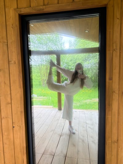 A young woman is captured in a dynamic pose, seemingly performing a split, as she takes a selfie reflected in a large, rectangular glass panel. The image is taken from above, looking down onto the window, which is framed by a black border and set into a light-colored wooden structure, possibly a floor or ceiling skylight in a building in Vozhoiskii, Russia. The wooden planks feature a natural grain.

The woman has long, light brown hair and is dressed in a light-colored, casual tracksuit or loungewear. She holds a silver smartphone in her right hand, pointed towards the glass to capture her reflection. Her pose is unusual, with one leg extended upwards in a split-like fashion and the other bent, creating an illusion that she is suspended or balancing. Her reflection appears to merge with the outdoor scenery.

Through the clear glass, a vibrant green natural environment is visible, featuring lush trees, grass, and scattered yellow flowers, indicative of a bright, daytime setting. A light-colored wooden deck or pathway can be seen immediately outside the window, where the woman's reflected leg seems to touch. The scene is well-lit, suggesting clear weather. No visible text is present in the image.