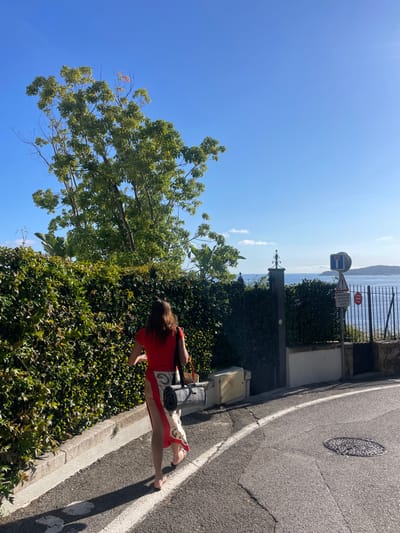 A female individual is depicted walking away from the camera on a paved road in Cap-d'Ail, France, under a clear blue sky. She wears a red short-sleeved top, a long beige and black patterned skirt, and light-colored flip-flop sandals. A dark shoulder bag is on her left shoulder, and a rolled-up dark grey mat, possibly a yoga mat, is slung over the same shoulder, resting against her left hip.

The road is asphalt, featuring a solid white line adjacent to a concrete curb on the right. A circular metal manhole cover with a textured pattern is visible on the road surface in the lower right quadrant. To the right of the road, a dense green hedge borders a property. Beyond the hedge, a dark metal gate with an ornate top is partially visible, alongside a light-colored utility box.

Further right, a metal pole supports two distinct road signs: a circular "No Entry" sign (red circle with white horizontal bar) and a triangular warning sign (red border, white background with a black exclamation mark). A smaller white rectangular sign with black text is mounted below these. Above the hedge and gate, the Mediterranean Sea is visible, extending to a distant landmass or island under the horizon. A large tree with green foliage dominates the upper left portion of the image. The scene is brightly illuminated by direct sunlight, casting long shadows to the left, indicating a mid-day or late afternoon setting.