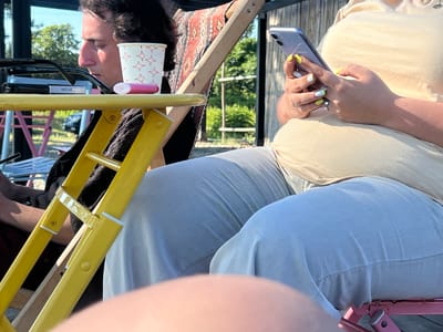 Two people are sitting outdoors on a sunny day, possibly in Ochamchire, Abkhazia. The person on the left, a man with dark, curly hair, is looking down at a yellow table. On the table, there's a white paper cup with red star-like patterns and a pink object that appears to be a vape pen. Next to it are a pair of dark-rimmed glasses and a cigarette pack labeled "MILAN". The man is wearing a dark shirt.

To the right, another person, whose face is not fully visible, is holding a smartphone in their hands and appears to be texting or browsing. This person is wearing a light beige top and light blue pants. Their fingernails are painted yellow and white. They are sitting in a deck chair with a patterned fabric. The background shows lush green trees and bamboo-like structures, suggesting a warm, possibly tropical or subtropical climate. The sun is casting strong shadows, indicating it is likely midday or late afternoon. The overall mood seems relaxed, with both individuals engaged in their own activities.