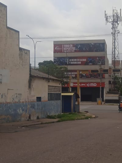 The image shows a street scene in Puerto La Cruz, Venezuela. In the background, a large building with several advertisements for motorcycle parts is visible. One advertisement reads "TODO EN REPUESTOS PARA MOTOS Y MAS" (Everything in spare parts for motorcycles and more). Another advertisement says "LO MEJOR EN CALIDAD Y DURABILIDAD" (The best in quality and durability). A cell tower is also visible in the background. In the foreground, a street with a faded blue and white wall on the left is shown. A small dog is walking on the sidewalk, and a motorcycle with an orange-clad rider is visible on the right side of the image. The sky is overcast.