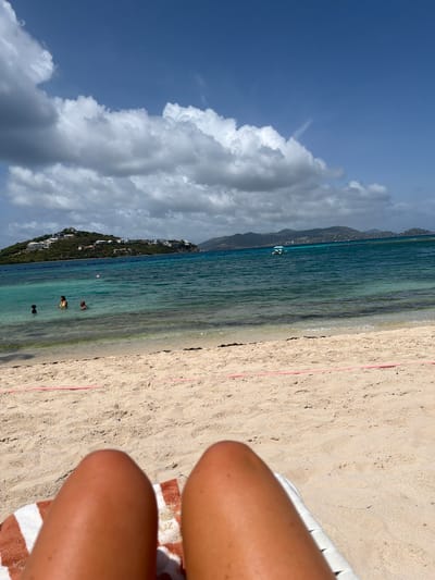 A first-person perspective image captures a beach scene in an unknown U.S. Virgin Islands location. The foreground is dominated by a sandy beach with a pink rope delineating an area. A portion of a towel and what appears to be legs are visible resting on a white lounge chair in the lower frame. The mid-ground shows the clear turquoise water of the ocean, with several people wading and swimming. Beyond the water, a lush green island rises, dotted with white buildings. The background features a mountainous landscape under a blue sky with scattered white and grey clouds. The overall scene depicts a sunny day at a tropical beach.