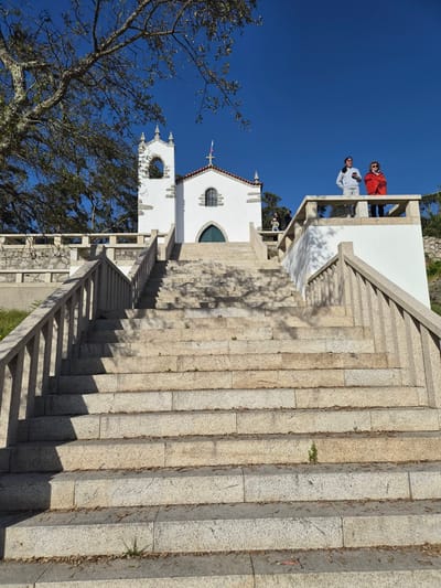 The image shows a long, broad stone staircase leading up to a white church building under a clear blue sky. Two people are standing on an upper balcony. A tree with bare branches is visible to the side of the church. The scene is located in Esposende, Portugal.