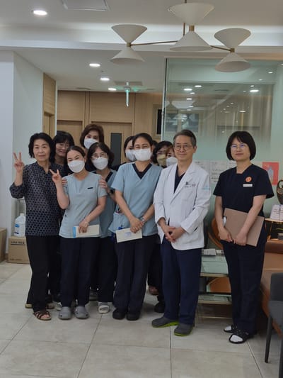 The image shows a group of nine individuals, appearing to be healthcare professionals, posing for a photo indoors. They are standing in what seems to be a clinic or hospital waiting area, characterized by tiled floors, light-colored walls, and modern lighting fixtures on the ceiling. In the background, there are doors, wooden paneling, and glass partitions.
To the far left, a woman wearing a dark patterned top and dark pants is making a peace sign with her left hand. Next to her are two other women, both wearing light blue scrubs and face masks. The woman in the center of this subgroup is also making a peace sign with her right hand and holding a white paper. To their right, a man in a white doctor's coat and dark pants stands with his hands clasped in front of him. He is wearing glasses and a face mask. Beside him is a woman in dark blue scrubs and glasses, also wearing a face mask and holding a light brown folder. The remaining individuals are partially obscured but appear to be women wearing similar scrub attire and face masks.
A white bottle of liquid, likely disinfectant, is visible on the floor to the left of the group. In the background, a table with chairs and a couch can be seen through a glass partition, suggesting a waiting or reception area.
The presence of "Suwon-si, South Korea" in the prompt implies that this scene is located in Suwon. Without further context or specific landmarks, pinpointing the exact location within Suwon is not possible from the image alone. However, the clean, modern interior suggests a healthcare facility.