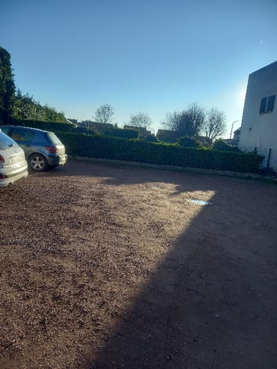 The image depicts an outdoor parking area in Noyelles-sous-Lens, France, under a clear, bright blue sky with a low sun. The foreground features a gravel and dirt surface, with a sharp, diagonal shadow line extending from the upper right to the lower left. A small puddle of water is visible within the sunlit portion of the gravel near the shadow line. Two passenger vehicles are parked on the left side: a light-colored hatchback, partially visible, and a darker Peugeot 307 hatchback, positioned further back and oriented away from the viewer. A dense, neatly trimmed green hedge forms a boundary across the midground. Behind the hedge, various trees with bare branches are visible, indicating a deciduous species, along with faint outlines of structures beyond. On the far right, a portion of a light-colored building with two dark, closed shutters on an upper floor is visible, partially obscured by direct sunlight creating lens flare. No people or active interactions are present in the scene.