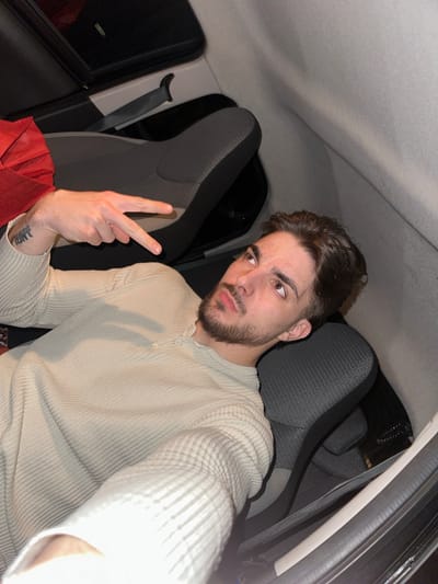 A person is inside a car, making a peace sign with their left hand. The person has brown hair and a beard, and is wearing a light-colored, long-sleeved shirt with buttons at the collar. A visible tattoo is on the person's left forearm. The car's interior consists of grey fabric seats and a dark-colored window frame. The environment suggests nighttime inside a car. There is no contextual information that suggests the location is Rafz, Switzerland.