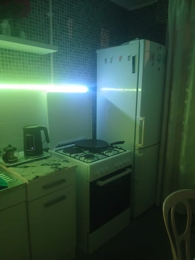 The image depicts a dimly lit kitchen with green and blue neon lighting. A white refrigerator stands on the right, adorned with several magnets. To its left, a white gas stove with a black stovetop is visible, featuring a black pan. In front of the stove, to the left, is a kitchen counter with a black electric kettle and a glass teapot on a tray. Underneath the counter are white cabinets with dark handles. The walls are tiled with small, dark squares. A light green curtain is visible to the right of the refrigerator. The floor is dark. A chair is partially visible in the bottom right corner. The scene is set in a domestic kitchen.